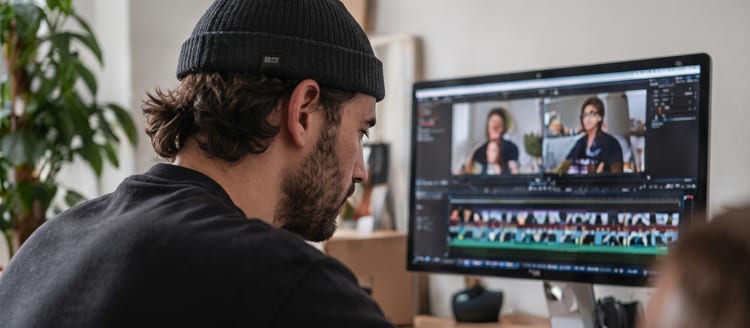 A man in a beanie editing video on a large monitor, seen over his shoulder