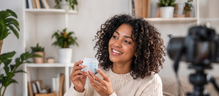 A woman with curly hair smiling as she holds up a small jar in front of a camera