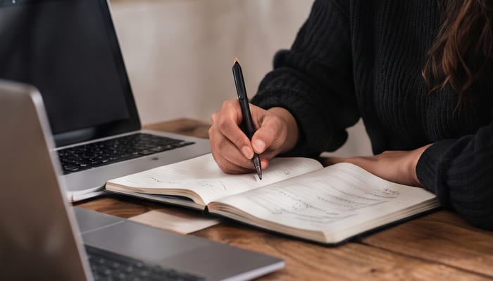 A hand writing in an open notebook on a wooden desk beside a laptop