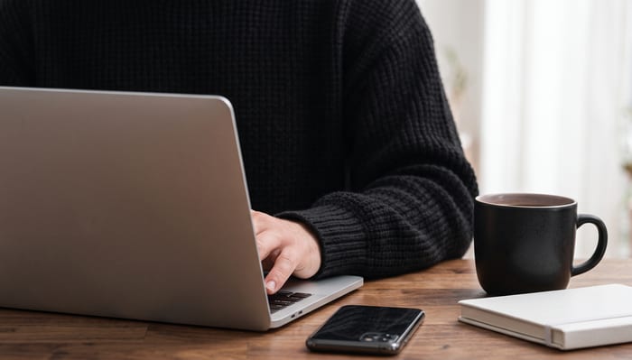 Hands typing on a laptop at a wooden desk beside a coffee mug and a phone